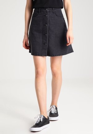 Femme portant une jupe en jean noire boutonnée à l'avant, associée à des baskets noires et blanches, debout sur un fond clair uni.