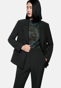 Schwarzer maßgeschneiderter Anzug mit strukturiertem Blazer und eng geschnittenen Hosen. Darunter ein gefaltetes Oberteil in dunklen Tönen mit grünen und blauen Akzenten.