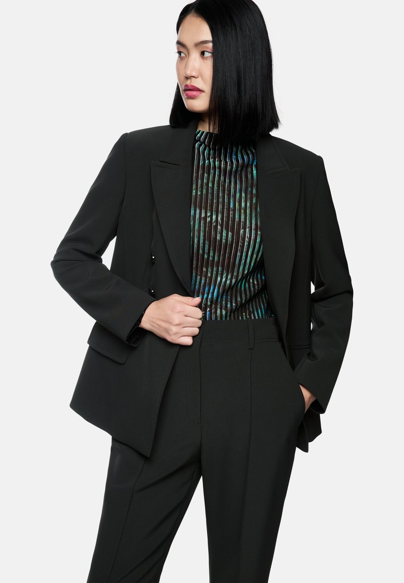 Schwarzer maßgeschneiderter Anzug mit strukturiertem Blazer und eng geschnittenen Hosen. Darunter ein gefaltetes Oberteil in dunklen Tönen mit grünen und blauen Akzenten.