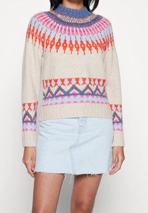 Kvinde iført en cremefarvet sweater med farverige geometriske mønstre i orange, pink, blå og en lyseblå denimnederdel med frynset kant.