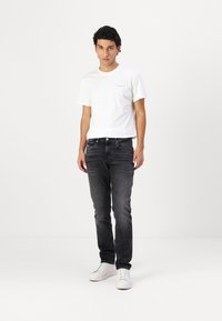 Weißes Baumwoll-T-Shirt mit kleinem Logo auf der Brust, kombiniert mit dunkelgrauen Jeans und weißen Sneakers. Einfaches und lässiges Outfit.