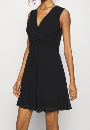 Femme portant une robe noire sans manches à col en V avec un détail froncé à la taille et un collier chaîne doré, debout devant un fond uni.