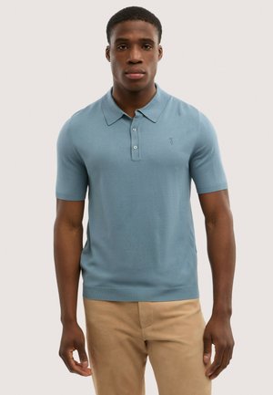 Giovane uomo che indossa una polo azzurra chiara a maniche corte aderente e pantaloni beige, in piedi contro uno sfondo chiaro e uniforme.