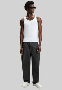 Débardeur blanc, pantalon large en denim foncé, baskets blanches. Le modèle porte des lunettes de soleil noires et se tient devant un fond gris.
