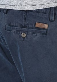 Námořnické modré bavlněné kalhoty s hladkou texturou, vybavené přední kapsou, knoflíkovým zapínáním a hnědou koženou nášivkou.