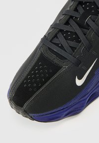 Μαύρο αθλητικό παπούτσι με υφή στην επιφάνεια, μεταλλικές λεπτομέρειες και μοβ σόλα. Διαθέτει διάτρητο σχέδιο και λογότυπο Nike σε λευκό χρώμα.