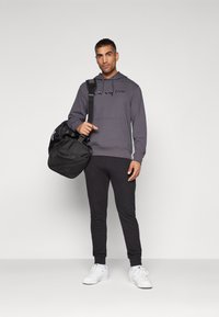 Sweat-shirt à capuche gris avec logo, joggeurs noirs et baskets blanches. Modèle tenant un sac de sport noir avec une bandoulière.
