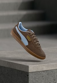 Bruna mockaskor med en ljusblå mockarand, rund tå, platta snören, gummisulor och en gul logotagg på sidan.