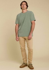 T-shirt vert olive à manches courtes associé à un jean skinny beige clair et des chaussures marron à semelle texturée, se tenant devant un arrière-plan neutre.