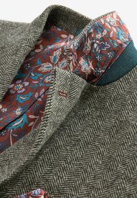 Blazer à motif chevron gris avec une doublure florale rouge et bleue. Dispose d'un revers cranté et d'accents en tissu contrastant.