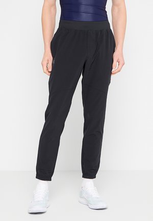 Personne portant un pantalon de sport noir avec des poignets élastiques et un haut sans manches bleu marine, debout sur un fond blanc avec des baskets blanches.
