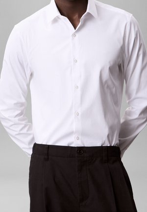 Homme portant une chemise blanche cintrée boutonnée, rentrée dans un pantalon noir taille haute, sur un fond neutre.