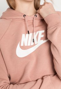 Rosa huvtröja med en vit Nike-logotyp, utrustad med en dragsko i huvan och svart hårdvara. Mjuk textur med en lös passform.