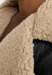 Giacca nera con ampio colletto in shearling beige soffice e spesso. Il tessuto strutturato contrasta con il materiale liscio nero sottostante.