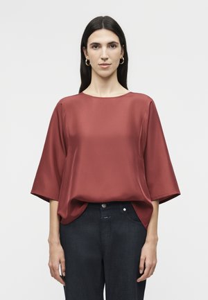 Donna con capelli lunghi e scuri indossa una camicetta di raso larga colore ruggine con maniche a gomito e pantaloni scuri a vita alta, in piedi.