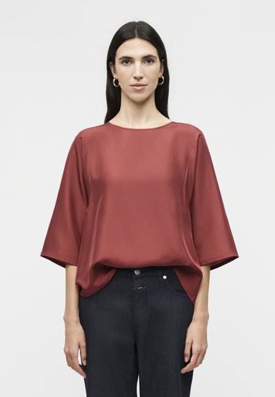 Woman with long dark hair wears a loose, rust-colored satin blouse with elbow-length sleeves and dark high-waisted pants, standing upright.