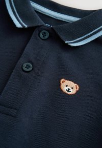 Tume sinine polo särk, mille krael on helesinised triibud. Kaks musta nööpi ja rinnal tikitud karu logo.