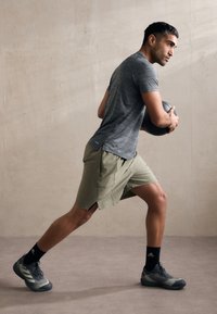Grå træningsshirt og olivenfarvede shorts med sidesplitter, sorte træningssko og en medicinbold, i en dynamisk træningsposition mod en neutral baggrund.