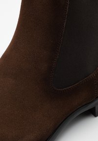 Bottine en daim marron avec panneaux latéraux élastiques, une texture lisse et un talon fin. Le design comprend des coutures contrastées pour plus de détails.