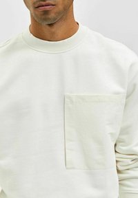 Ljuskrämfärgad sweatshirt med rund hals, textilstruktur och en enda fyrkantig bröstficka. Ren design med minimal detaljering.