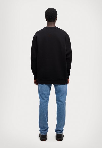 Sudadera negra de gran tamaño con puños y dobladillo acanalados, combinada con jeans de mezclilla azul claro y zapatillas negras, vista desde la parte posterior.