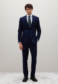 Abito navy con revers a punta, bottone singolo e due tasche frontali. Abbinato a una camicia bianca e cravatta verde a righe. Scarpe nere.