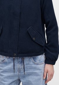 Personne portant une veste côtelée bleu foncé avec des poches à boutons-pression et un jean en denim bleu clair avec des cordons à la taille.