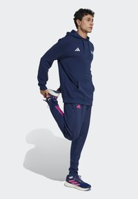 Dunkelblaue Kapuzenjacke und Jogginghose, beide mit rosa Akzenten. Die Kapuzenjacke verfügt über einen Kordelzug, eine Kängurutasche und ein Logo. Sportliche Schuhe mit Logo.