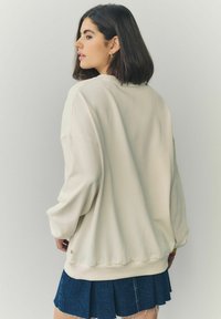 Béžová oversized mikina s okrúhlym výstrihom a spustenými plecami, kombinovaná s tmavomodrou skladanou sukňou vyrobenou z denimu. Hladká textúra látky.