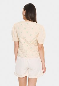 Quiltani prsluk u svijetlopeteljastoj boji sa zelenim uzorcima srca, kratkim napuhanim rukavima i okruglim izrezom, u kombinaciji s bijelim šortsevima.