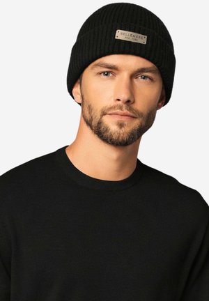 Mann mit leichtem Bart, der eine schwarze, gerippte Strickmütze mit der Aufschrift "Bellemere New York" und einen schwarzen Pullover mit Rundhalsausschnitt trägt.