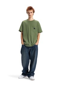 Olivově zelené tričko s kapsou na prsou, v kombinaci s volnými tmavě modrými džíny a bílými teniskami, stojící na bílém pozadí.