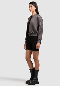 Kvinde med langt mørkt hår iført en grå satin bomberjakke, sort cropped top, sorte cykelshorts og sorte snørelåste kampstøvler.
