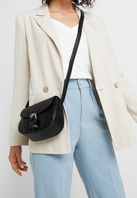 Žena v béžovém saku, bílém topu a světle modrých kalhotách s vysokým pasem, která nese černou koženou crossbody tašku s kovovou přezkou.