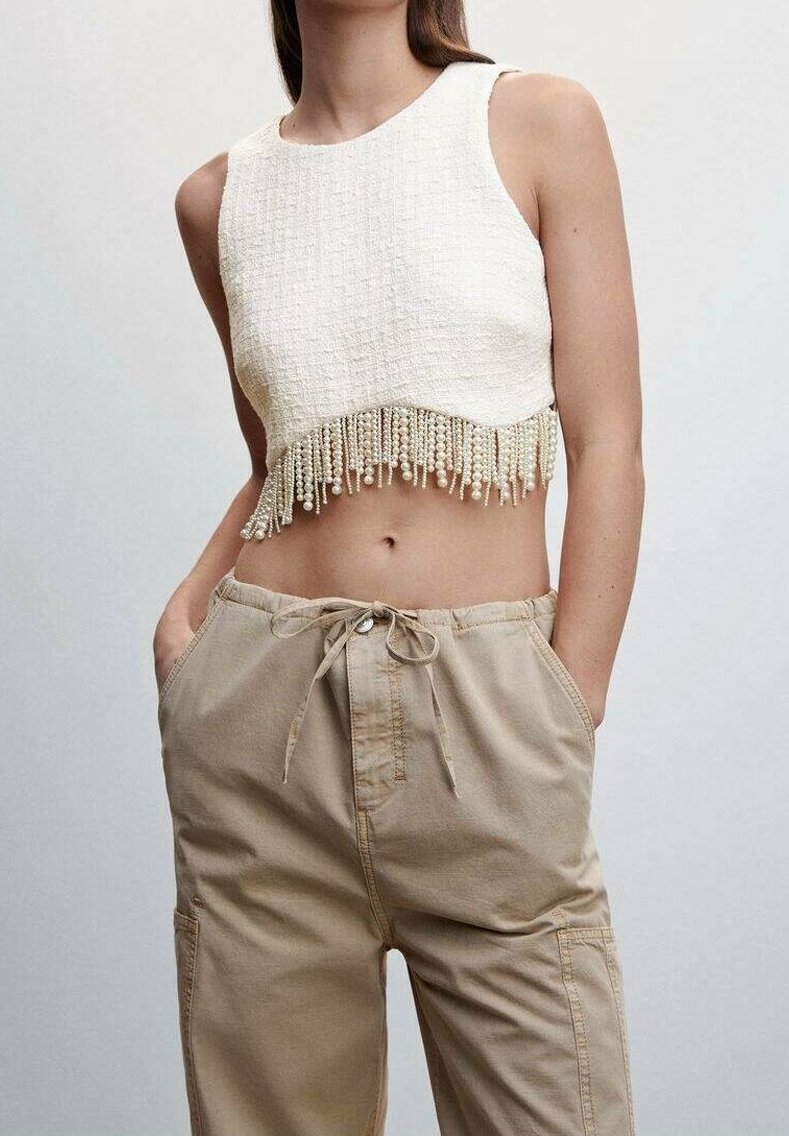 Top court sans manches blanc avec des détails en franges de perles, associé  à un pantalon cargo taille haute beige avec cordon de serrage, les mains dans les poches.