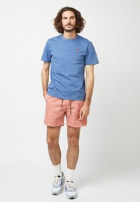 Maglietta di cotone blu con un piccolo logo rosso, abbinata a pantaloni corti rosa e calze bianche. L'outfit include scarpe da ginnastica con dettagli azzurri.