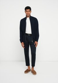 Giacca navy con zip, maglietta bianca a girocollo, pantaloni navy su misura, scarpe slip-on marroni con dettagli in espadrille, design minimalista.