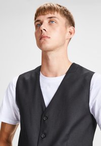 Chaleco negro con una textura suave, con un diseño de cuello en V y tres botones. Llevado sobre una camiseta blanca, diseñado para un ajuste entallado.