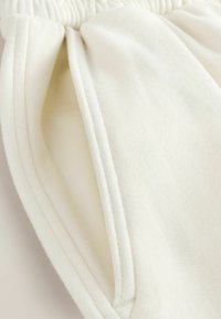 Primo piano di tessuto color crema con vita elastica e tasca laterale cucita su abbigliamento morbido.