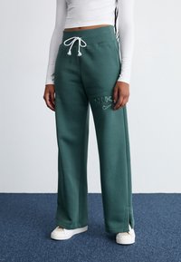 Zelené tepláky z měkké látky, s nastavitelným pasem na šňůrku, bočními rozparky a vyšitým logem „NIKE“ s malým detailem ve tvaru swooshe.