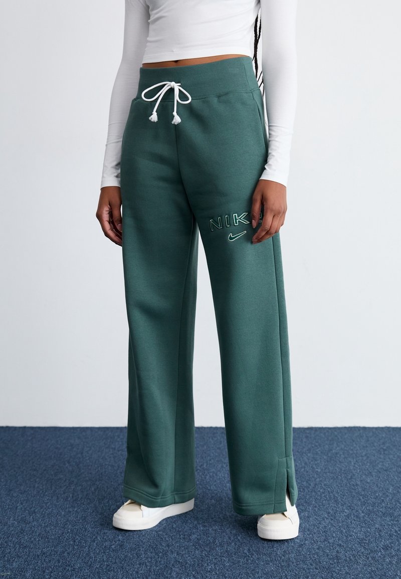 Zelené tepláky z měkké látky, s nastavitelným pasem na šňůrku, bočními rozparky a vyšitým logem „NIKE“ s malým detailem ve tvaru swooshe.