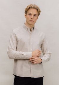 Lys beige skjorte med knappelukking i bomullsstoff med lange ermer, krage og hvite knapper; har rett kant og minimal tekstur.