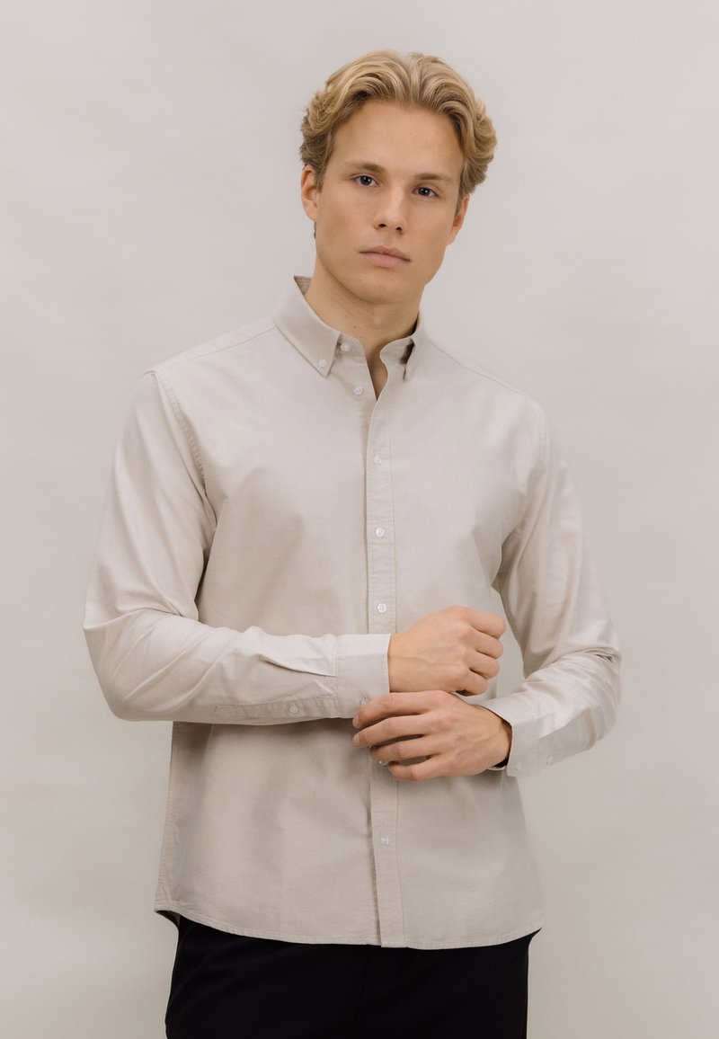 Lys beige skjorte med knappelukking i bomullsstoff med lange ermer, krage og hvite knapper; har rett kant og minimal tekstur.