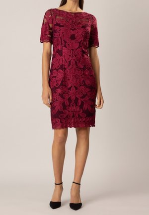 Kobieta w bordowej sukience za kolano z krótkim rękawem, ozdobionej misternymi kwiatowymi wzorami z koronki, oraz w czarnych butach na obcasie z paskiem wokół kostki, stojąca na jednolitym tle.