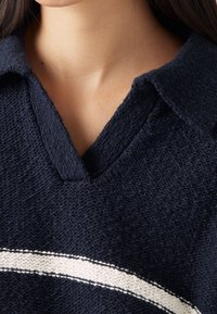 Námořnický pletený svetr s výstřihem do V a bílým vodorovným pruhem přes hrudník. Má texturovanou látku a strukturovaný límec.