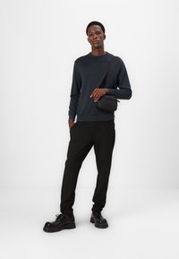 Marineblauer Pullover, schwarze Nadelstreifenhose und schwarze klobige Schuhe. Eine kleine schwarze Umhängetasche hängt über der Schulter. Schlichter Hintergrund.