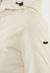 Giacca color crema con una consistenza liscia, dotata di cappuccio regolabile e un piccolo logo nero sulla manica.