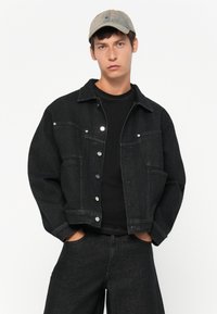 Young man wearing a faded gray cap, black denim jacket with silver buttons, black shirt, and black pants, standing with hands in pockets.