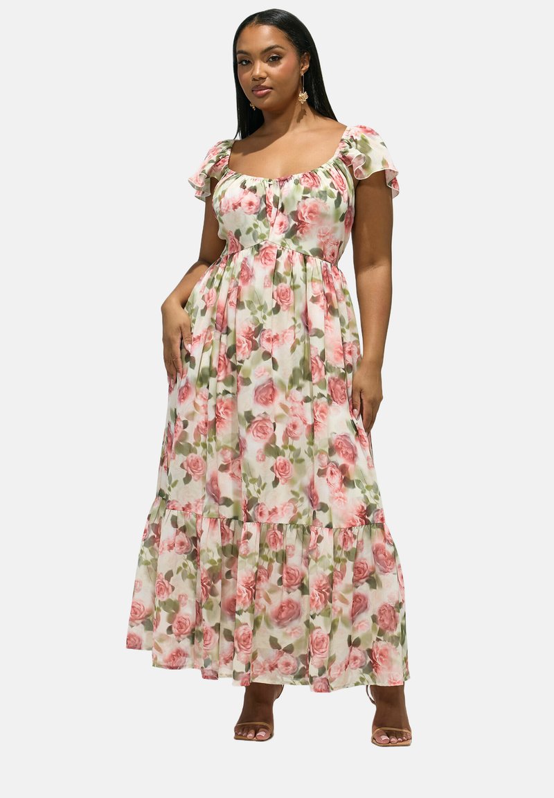 Frau trägt ein langes, fließendes, schulterfreies Kleid mit pinkfarbenen Rosenmustern und kurzen, gerafften Ärmeln, steht vor einem schlichten Hintergrund.