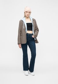 Νεαρή γυναίκα που φοράει μπεζ σκουφάκι, καφέ καρό σακάκι πάνω από ανοιχτό μπλε φούτερ με κουκούλα, μαύρο crop top, σκούρο μπλε τζιν και λευκά αθλητικά παπούτσια, στέκεται μπροστά σε λευκό φόντο.